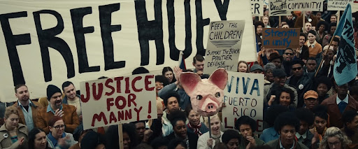This image shows a large, diverse group of people at a protest. Several are holding signs, which read "Free Huey," "Justice for Manny!," "Viva Puerto Rico," "Feed the Children," and other messages. In the foreground of the photo is a representation of a pig head on a stick.