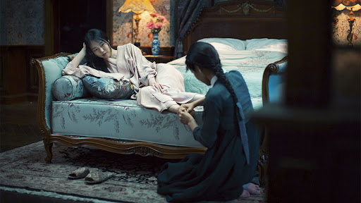 In this image, a woman in a pink robe is lounging while another woman kneels and massages her feet. We can consider the different positions and actions of the two women to reveal differences between their class and wealth statuses.